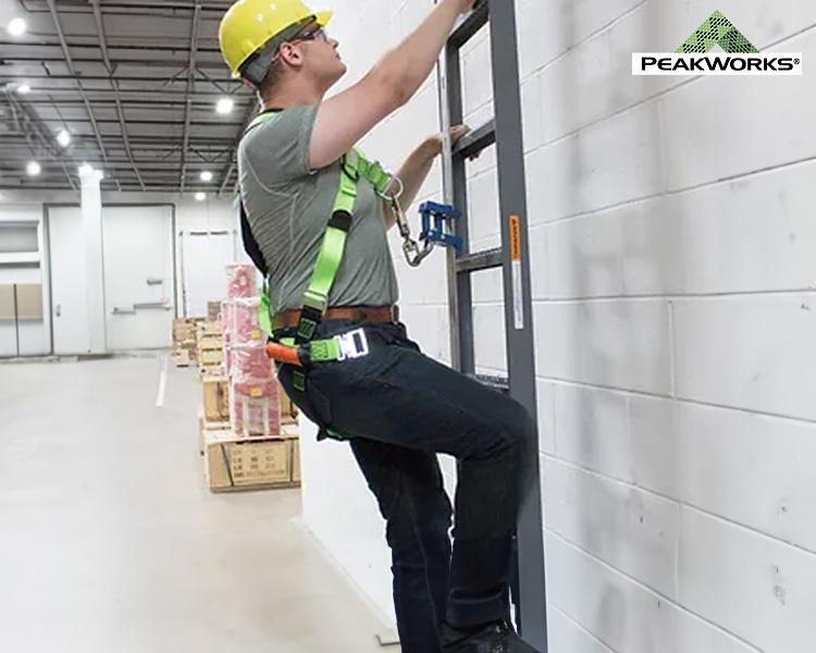 Construction worker in Peakworks yellow hard hat and green safety harness installing equipment on a wall, showcasing safety in a warehouse environment